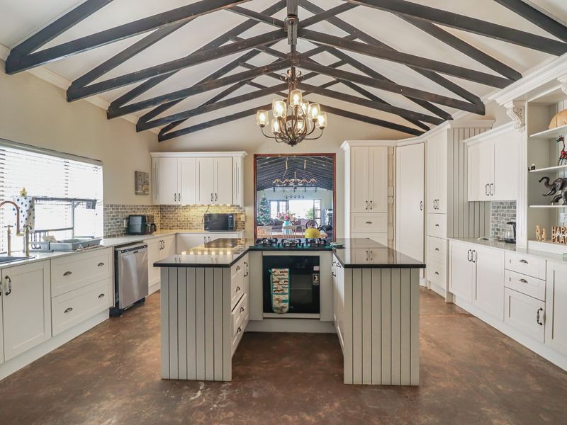 Spacious farmhouse kitchen featuring exposed timber trusses, a central island with built-in oven and hob, shaker cabinetry, and durable concrete flooring.