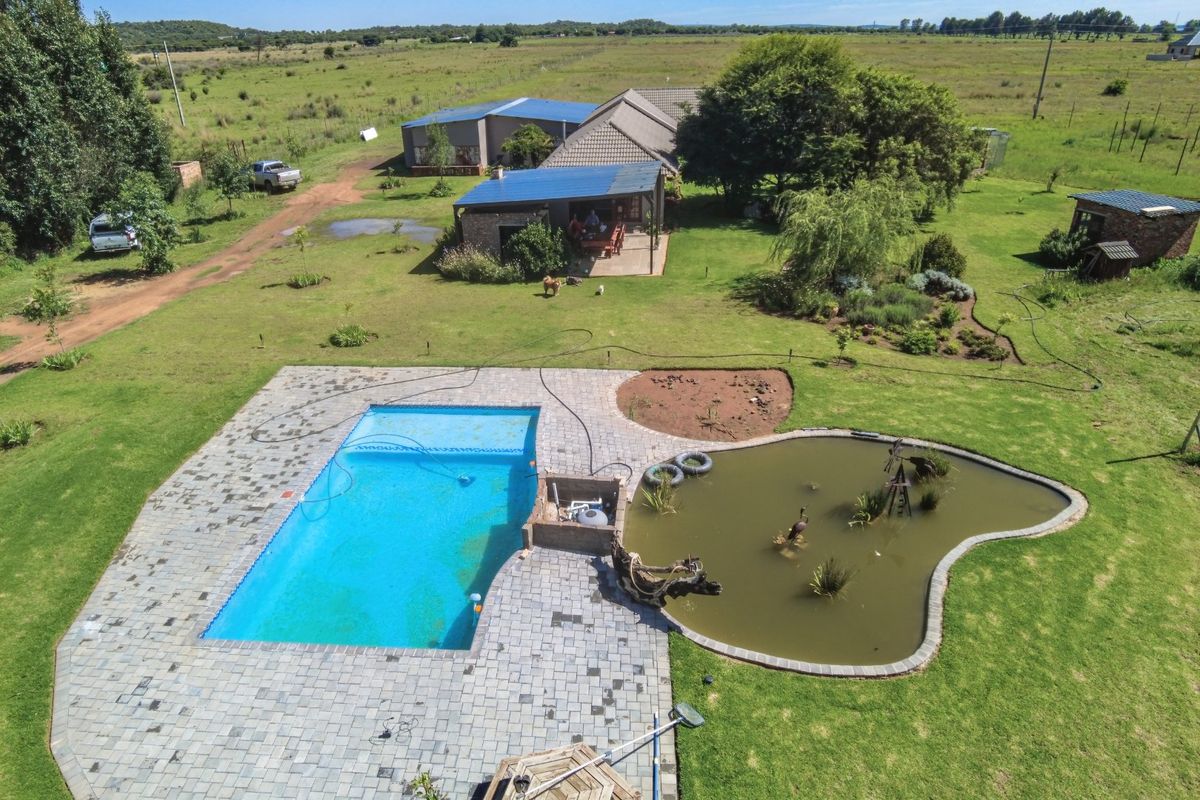 Aerial view of a farm property featuring a swimming pool, a natural pond, a paved patio area, and surrounding landscaped grounds.