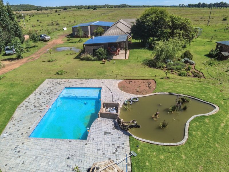 Aerial view of a farm property featuring a swimming pool, a natural pond, a paved patio area, and surrounding landscaped grounds.