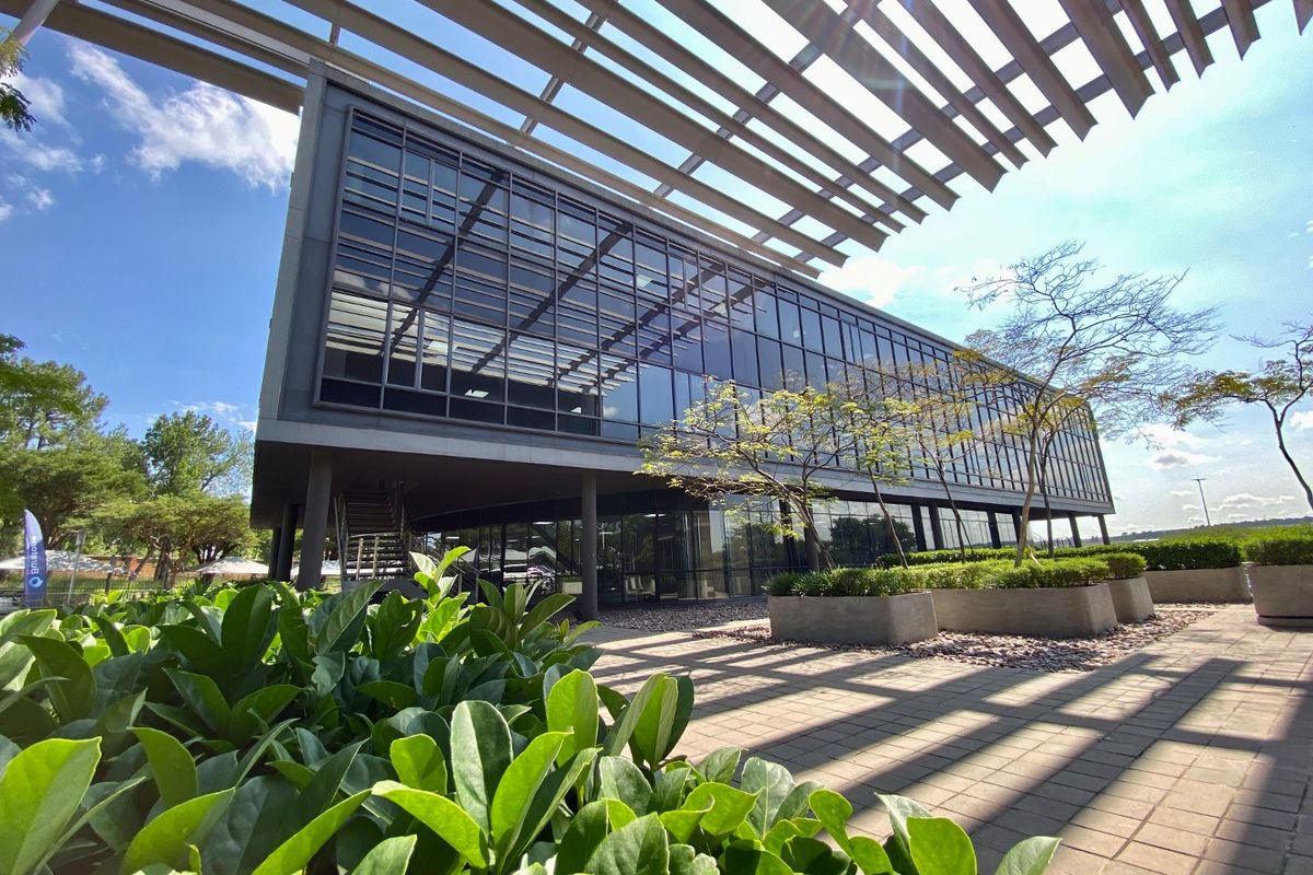 Modern commercial building featuring extensive glass curtain walling, a prominent louvred roof canopy, and a landscaped paved terrace area.