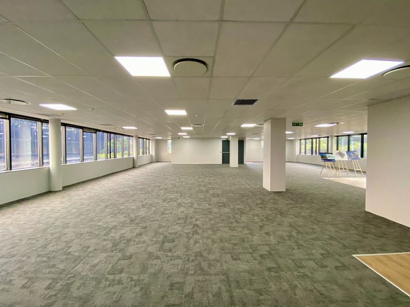 A spacious, open-plan commercial office area featuring neutral carpet tiles, a suspended ceiling with recessed lighting, and large windows providing ample natural light.