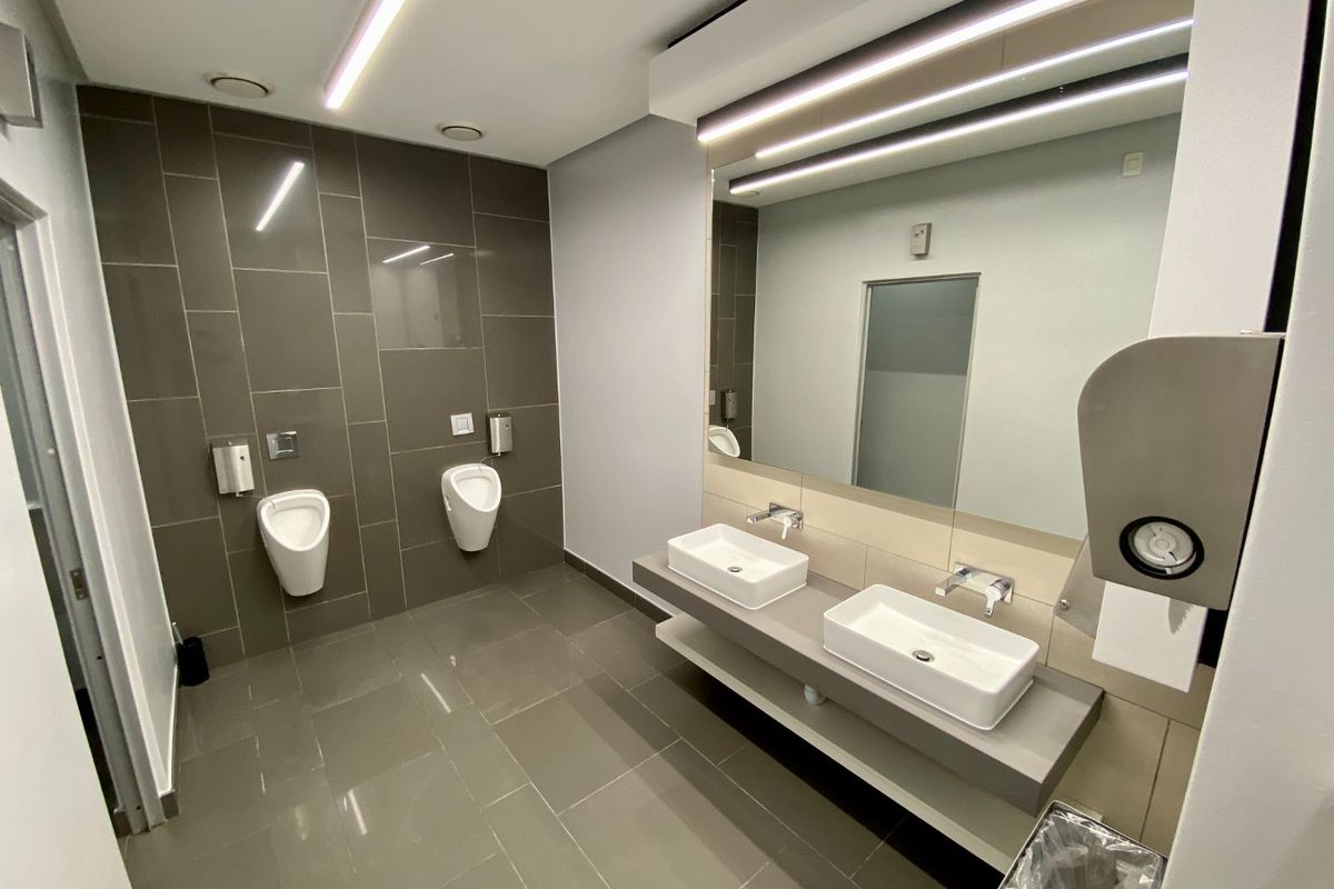 Modern commercial bathroom featuring dual vessel sinks, wall-mounted urinals, large mirror, and contemporary tiled finishes with integrated lighting.