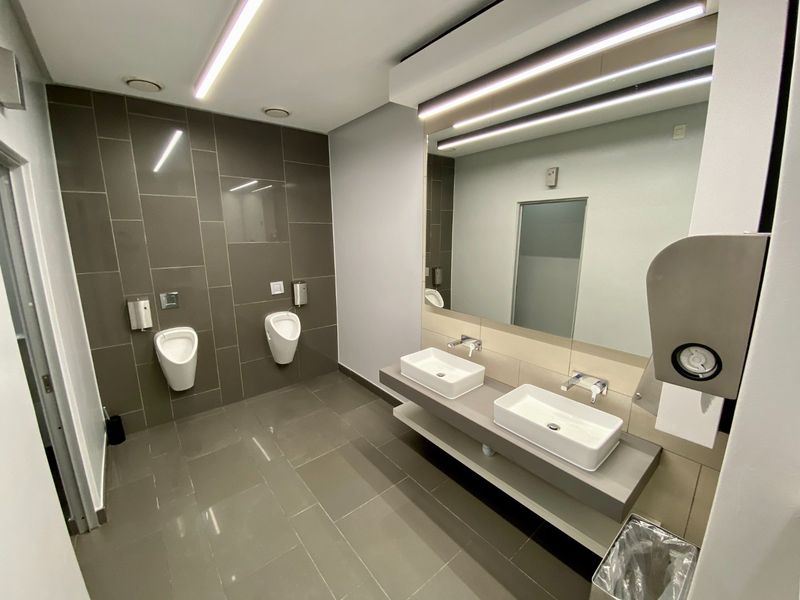 Modern commercial bathroom featuring dual vessel sinks, wall-mounted urinals, large mirror, and contemporary tiled finishes with integrated lighting.