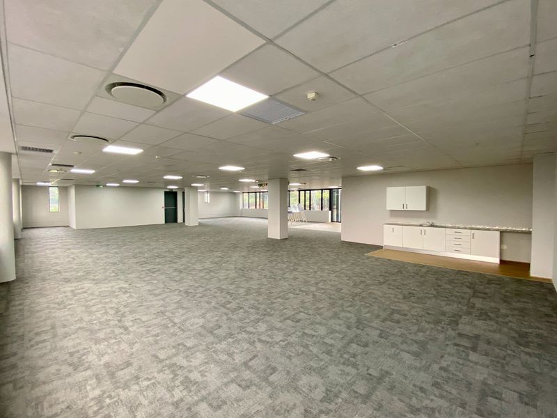 Spacious open-plan commercial office area featuring a suspended ceiling, recessed lighting, grey carpeting, and a compact built-in kitchenette unit.