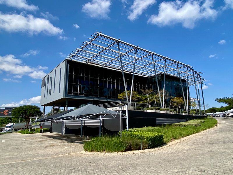 Modern commercial building featuring an elevated glass facade, extensive steel pergola structure, and integrated paved parking with landscaped garden surrounds.