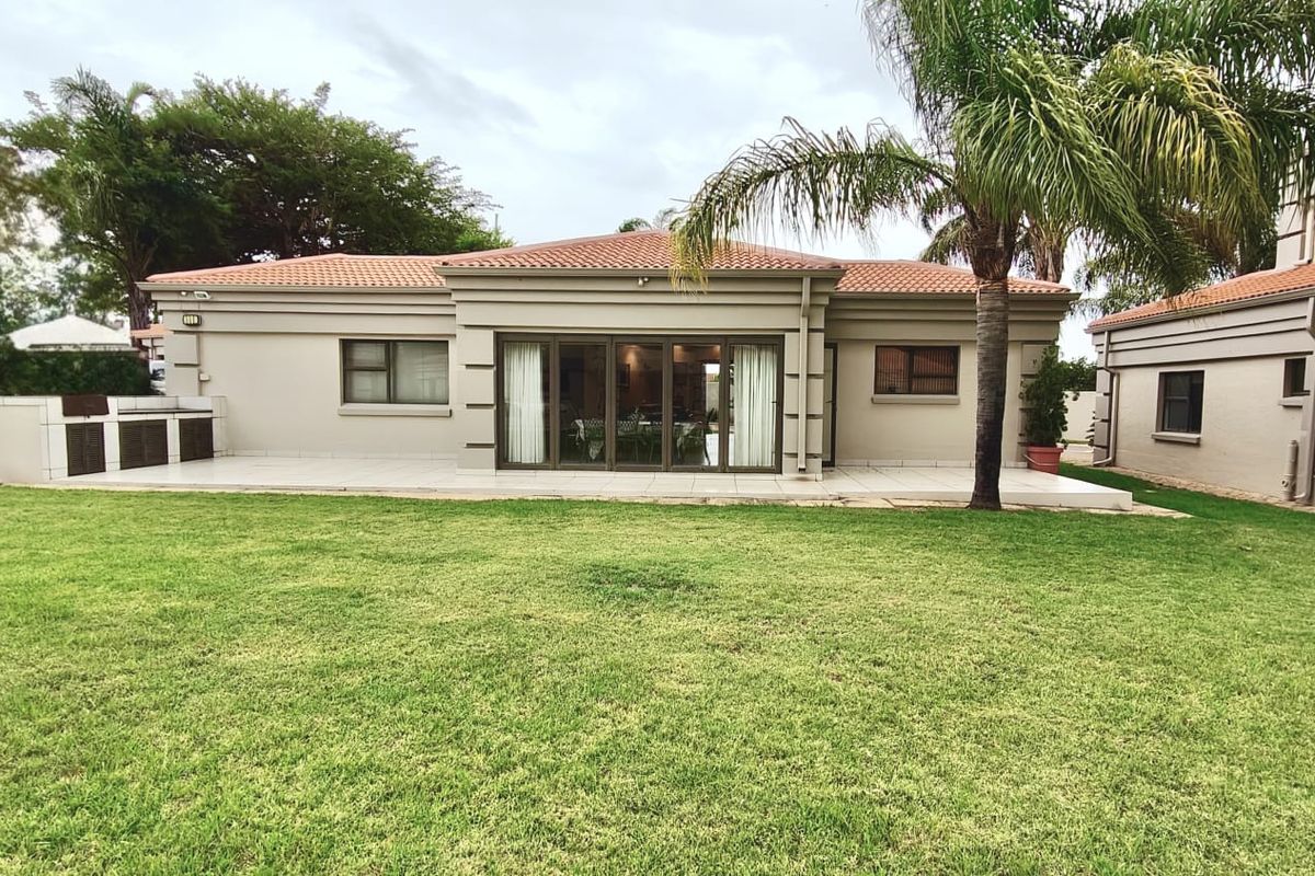 Rear view of the property featuring a spacious lawn, a tiled patio area with a built-in braai, and large sliding doors leading indoors.