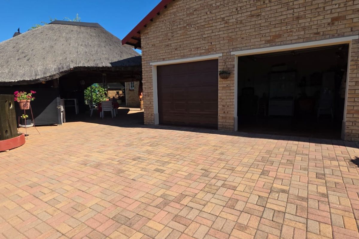 A paved driveway area leading to a double garage, featuring a thatched lapa structure in the background.