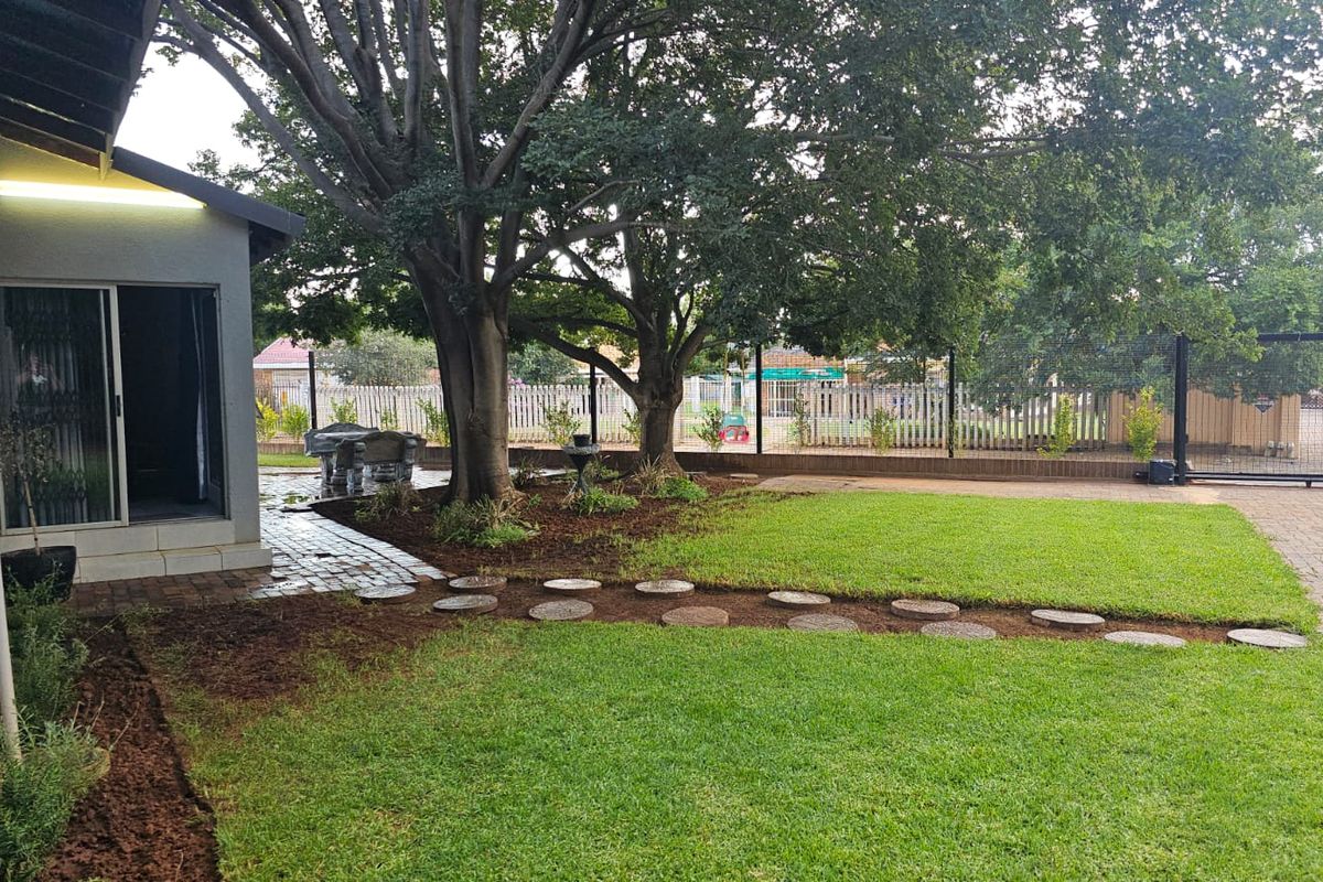A well-maintained rear garden featuring a manicured lawn, paved pathways, mature trees, and secure perimeter fencing with a sliding gate.