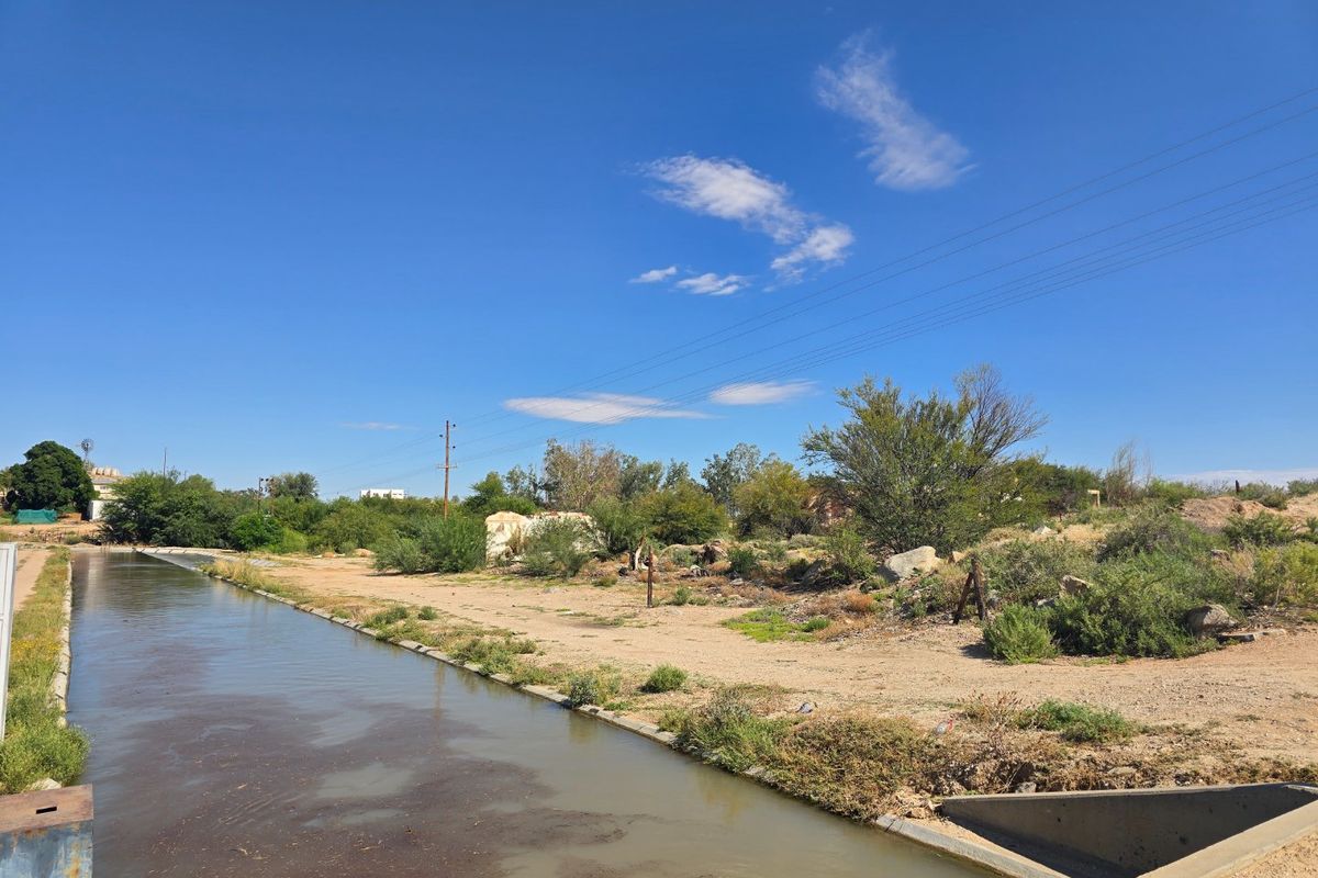 An outdoor view of a concrete irrigation canal bordering an undeveloped, overgrown plot of land with visible power lines.