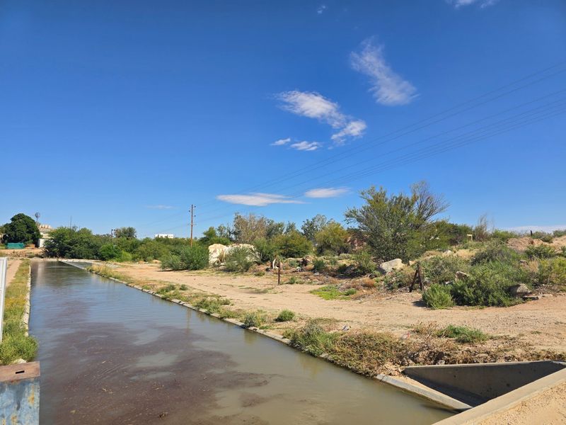 An outdoor view of a concrete irrigation canal bordering an undeveloped, overgrown plot of land with visible power lines.