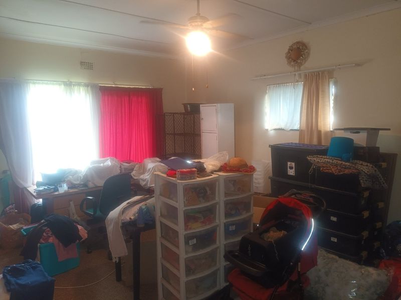 A bedroom space featuring a ceiling fan and ventilation grille, currently used for storage with significant clutter throughout the room.