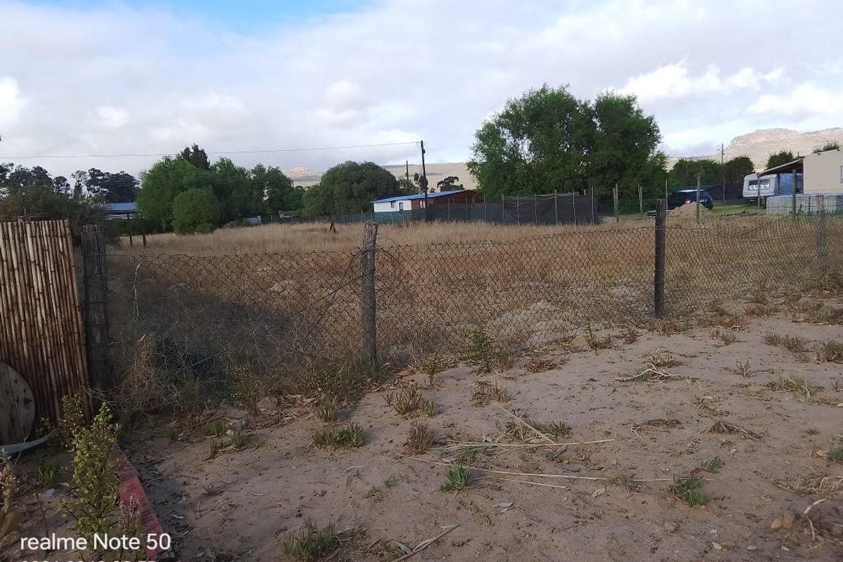 A view of a vacant residential plot featuring sandy ground, overgrown grass, and perimeter chain-link fencing.