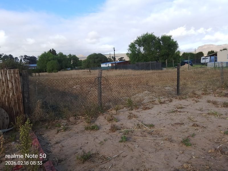 A view of a vacant residential plot featuring sandy ground, overgrown grass, and perimeter chain-link fencing.