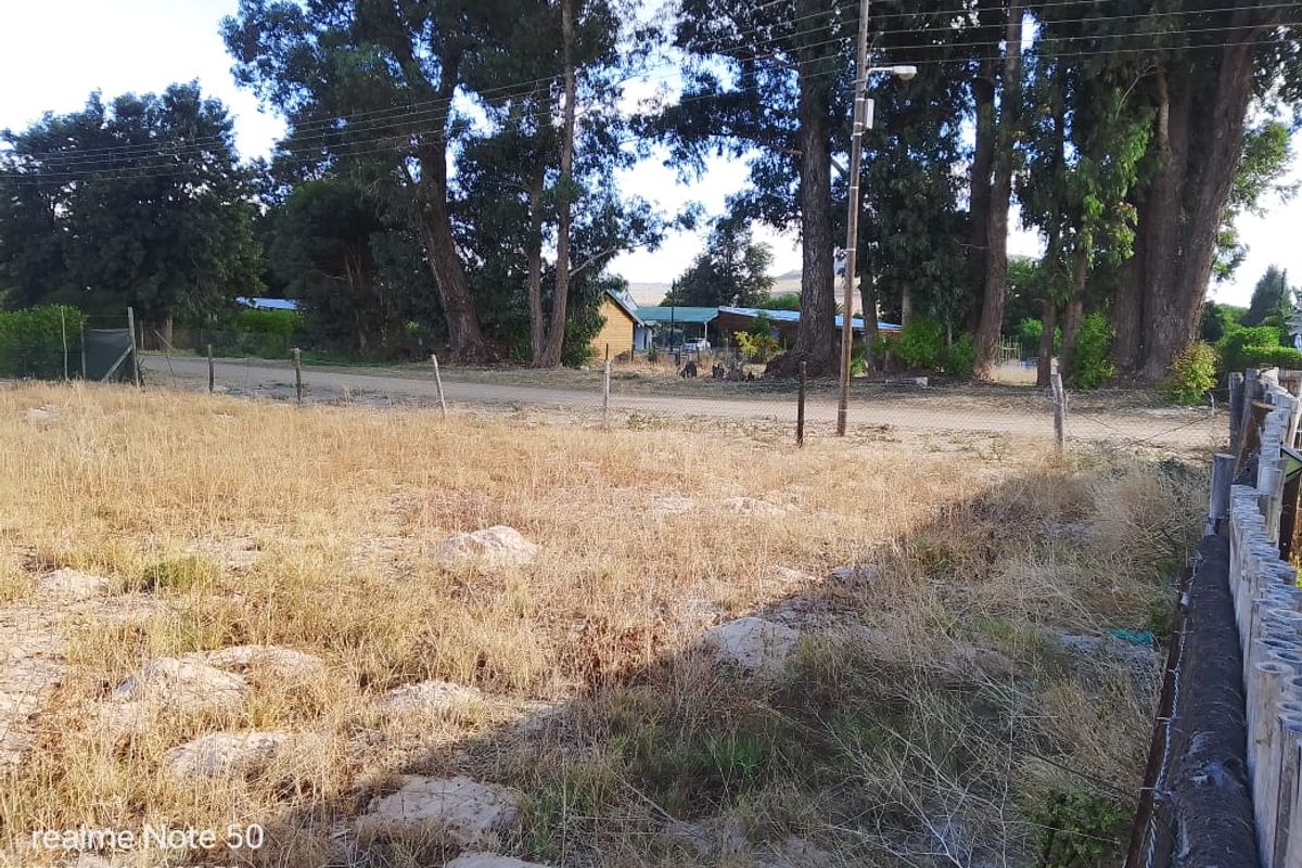 A view of a vacant residential plot featuring dry grass, mature trees, and perimeter wire fencing along a dirt road.