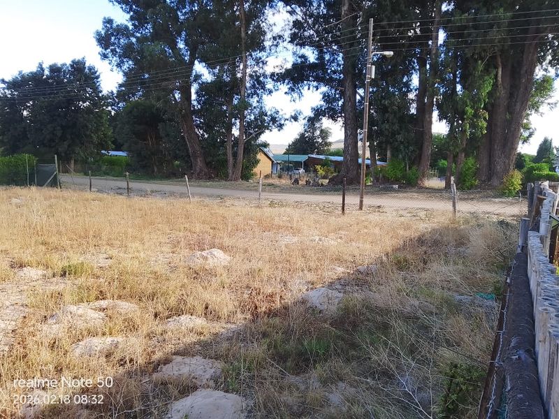 A view of a vacant residential plot featuring dry grass, mature trees, and perimeter wire fencing along a dirt road.