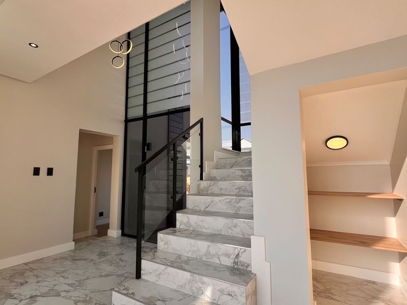 Modern double-volume entrance hall featuring a striking marble-look staircase with glass balustrades, high windows, and convenient built-in under-stair storage shelving.