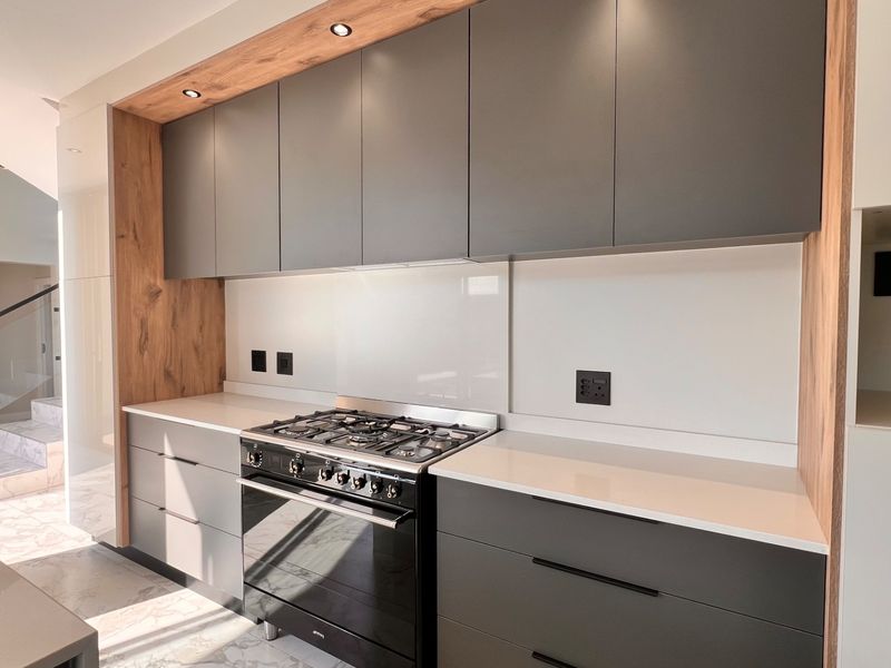 Modern kitchen featuring sleek grey cabinetry, wood-grain accents, quartz countertops, and a high-end gas stove with a glass splashback.