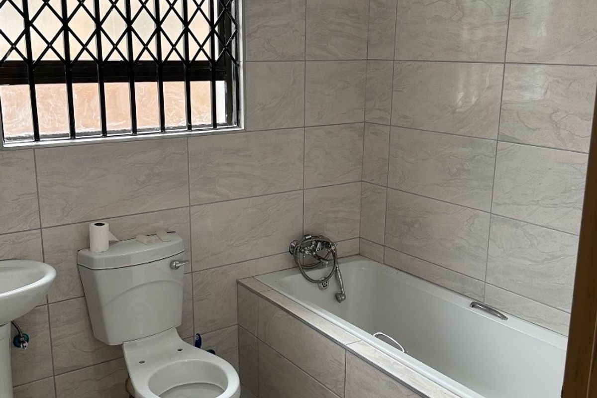 A functional bathroom featuring full-height wall tiling, a pedestal basin, a built-in bathtub with mixer tap, and a window with a security gate.