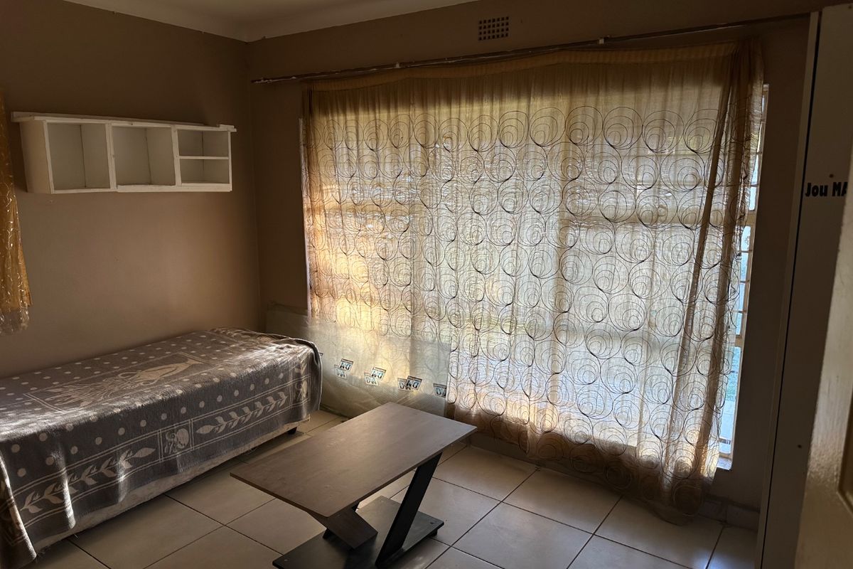 A bedroom featuring tiled flooring, built-in wall shelving, and a large window with security bars, offering a functional space with potential for cosmetic updates.