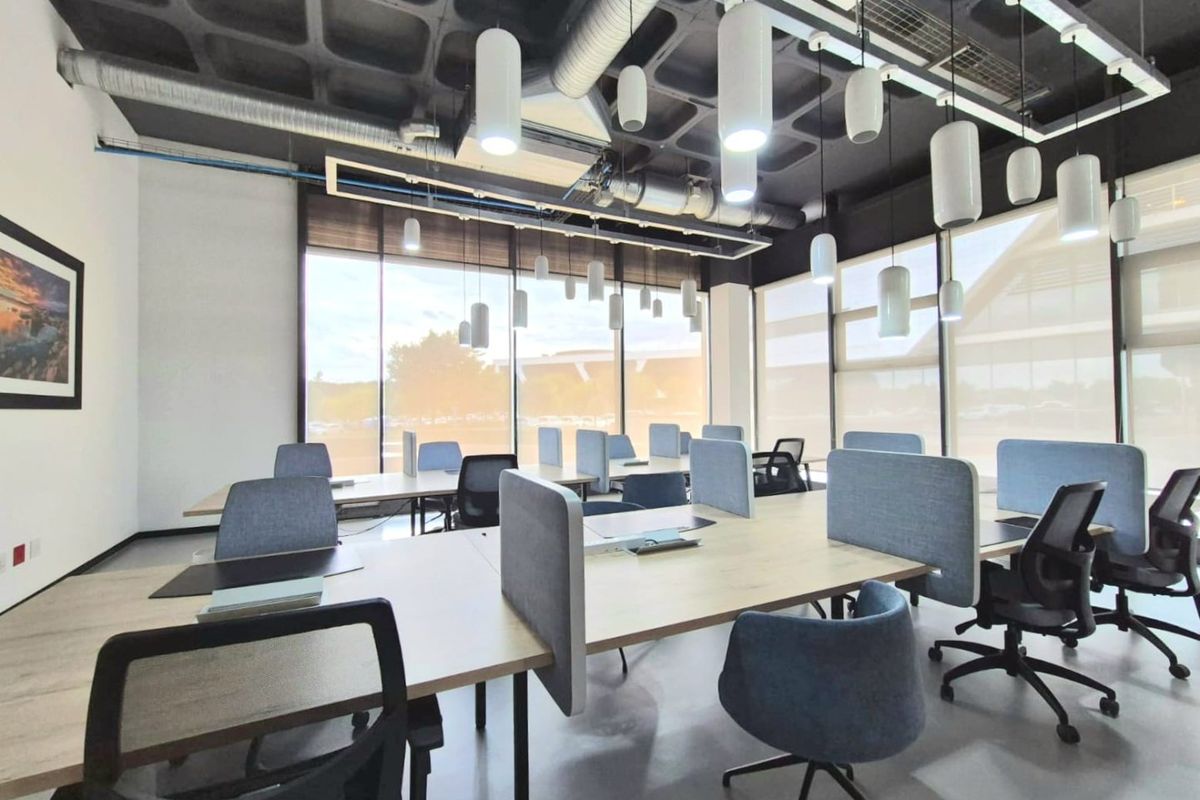Modern open-plan office space featuring an exposed waffle slab ceiling, industrial ducting, large windows with roller blinds, and contemporary pendant lighting.