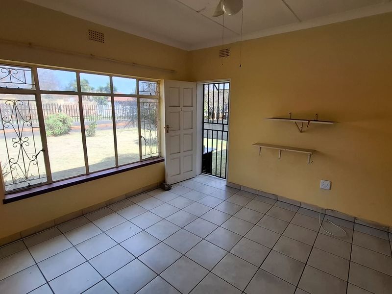 An empty living room featuring tiled flooring, a large window with security bars, and direct access to the garden through a security gate.