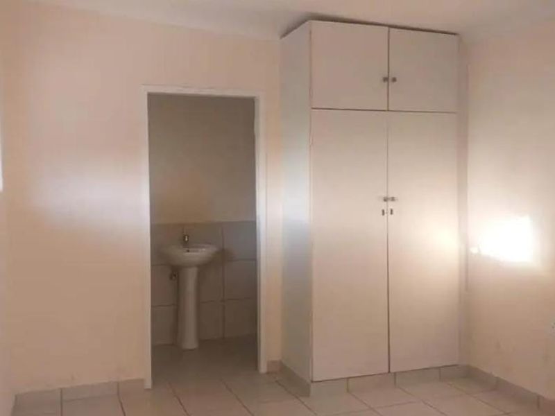 A simple bedroom featuring tiled flooring, a built-in wardrobe, and an en-suite area with a pedestal basin.