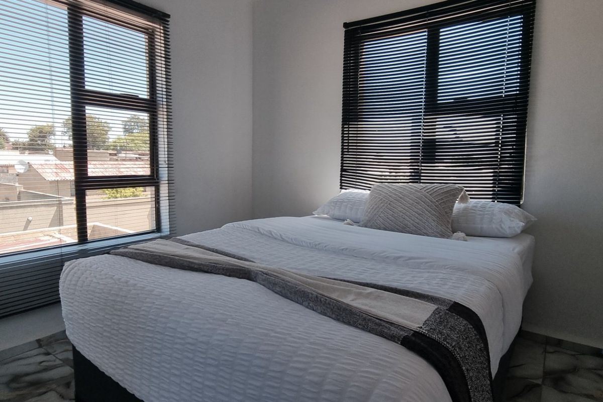 A bright bedroom featuring tiled flooring, large windows with fitted venetian blinds, and neutral wall finishes.