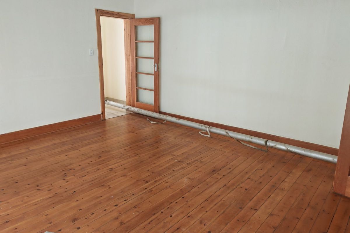 An empty living room featuring original timber flooring and a timber door, currently obstructed by exposed pipework running across the floor.