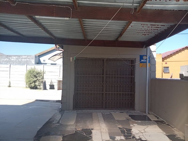 A covered parking area or garage featuring a corrugated metal roof, security gate, and paved flooring, situated within a fenced property.