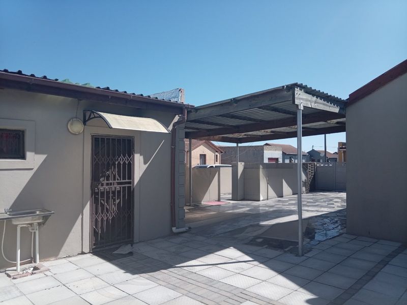 A paved outdoor patio area featuring a covered carport structure, security gate, and an external wash basin, providing functional space for parking or outdoor activities.