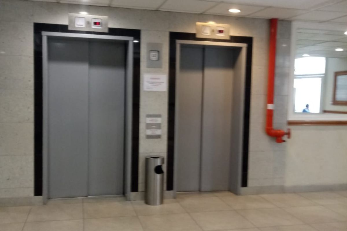 A communal apartment building hallway featuring two elevator doors, tiled flooring, and a suspended ceiling.