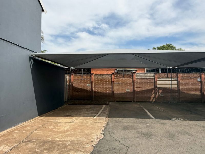 Outdoor parking area featuring a shade cloth carport structure, asphalt paving, and a secure brick boundary wall with razor wire.