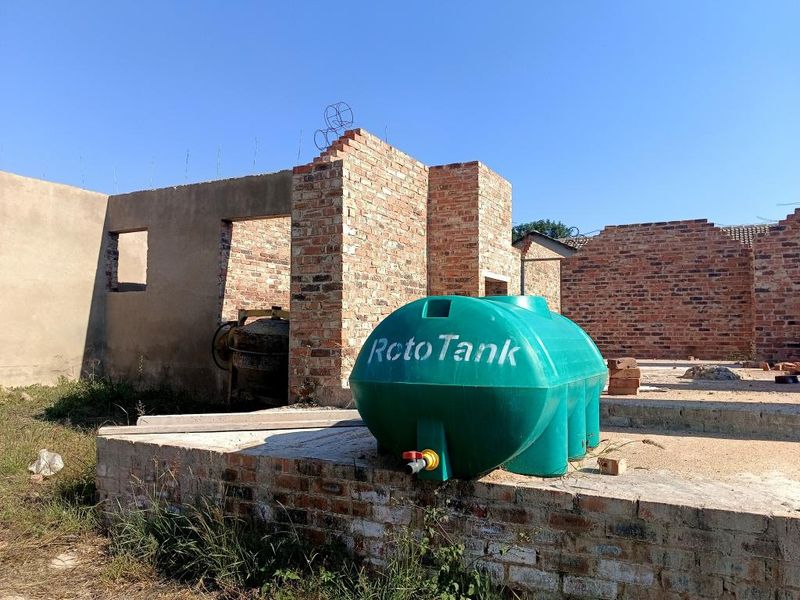 An unfinished residential construction site showing exposed brick walls, a concrete foundation slab, and a plastic water storage tank on the property.
