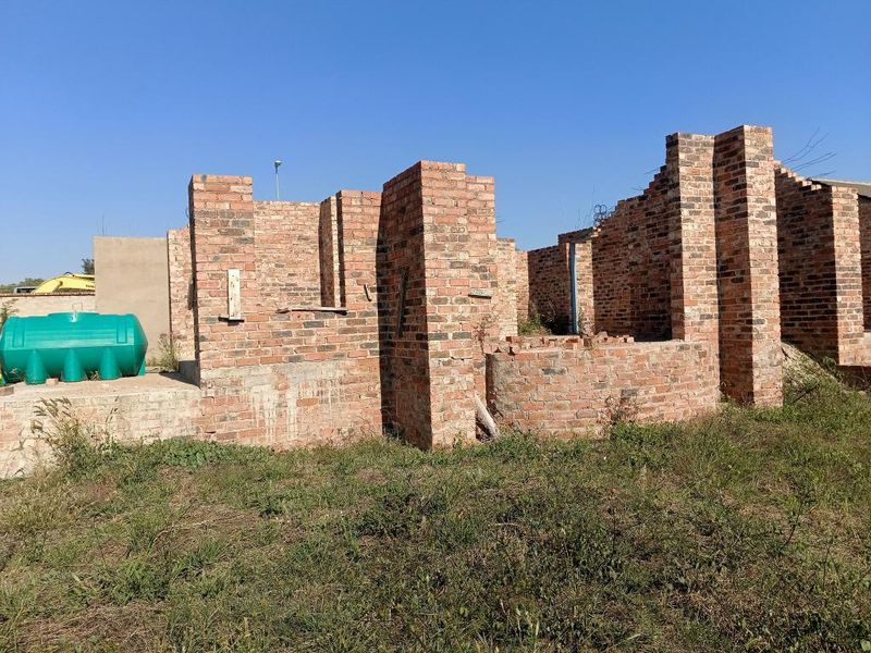 An unfinished residential construction site showing exposed brick walls and a water storage tank on a vacant plot.