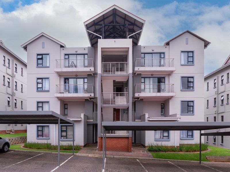 Front elevation of a modern multi-story apartment complex featuring private balconies, covered parking bays, and a central stairwell entrance.