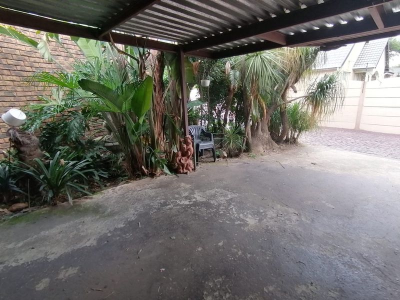 A covered outdoor parking area featuring a corrugated metal roof and a concrete driveway, bordered by established garden foliage and a brick wall.