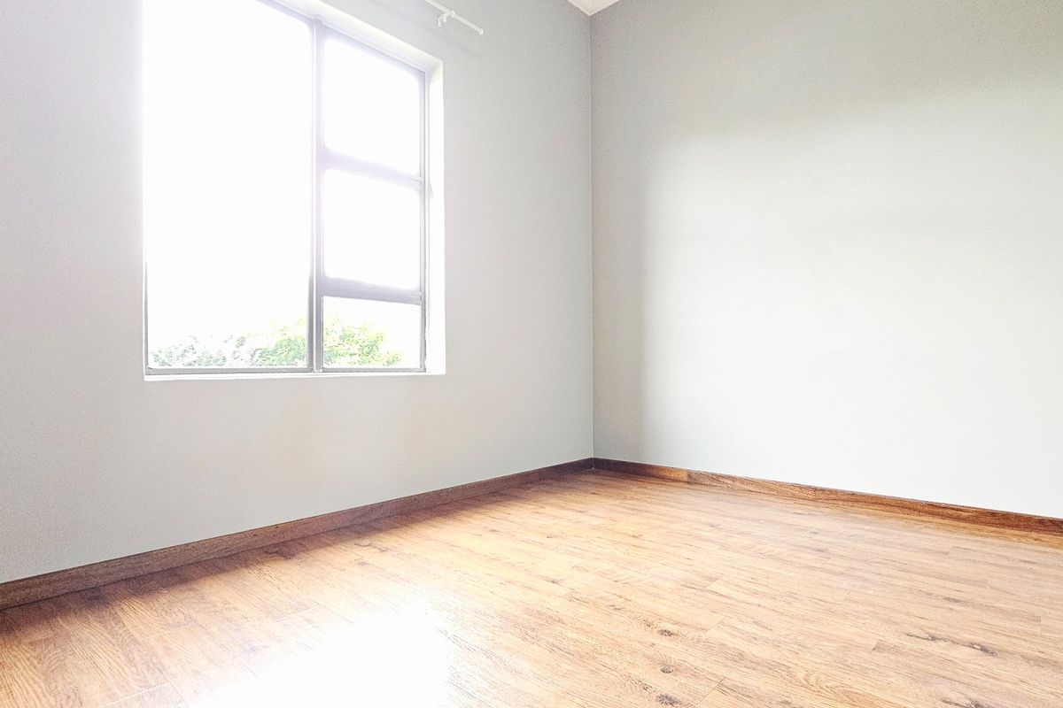 A bright, empty bedroom featuring neutral walls, laminate flooring, and a large window providing natural light.