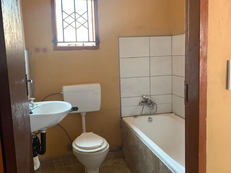 A compact bathroom featuring a bathtub, pedestal basin, and toilet, with tiled splashbacks and a security-grille window. Requires cosmetic updates.