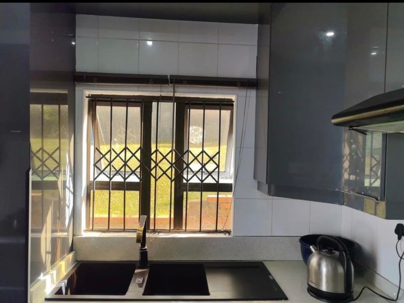 Modern kitchen area featuring dark cabinetry, stone countertops, a double basin sink, and a window fitted with security bars overlooking the garden.