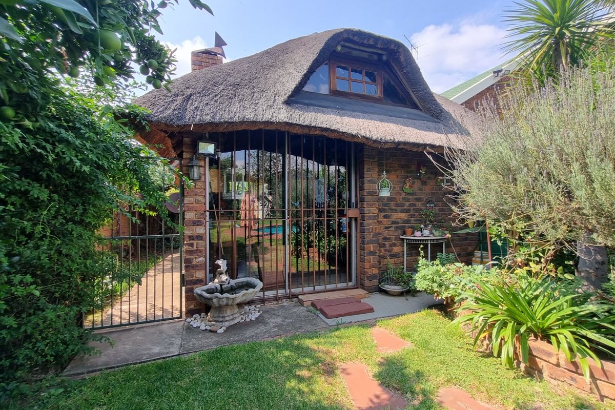 A charming facebrick cottage featuring a traditional thatch roof, security gate, and sliding glass door, set within a lush, established garden.