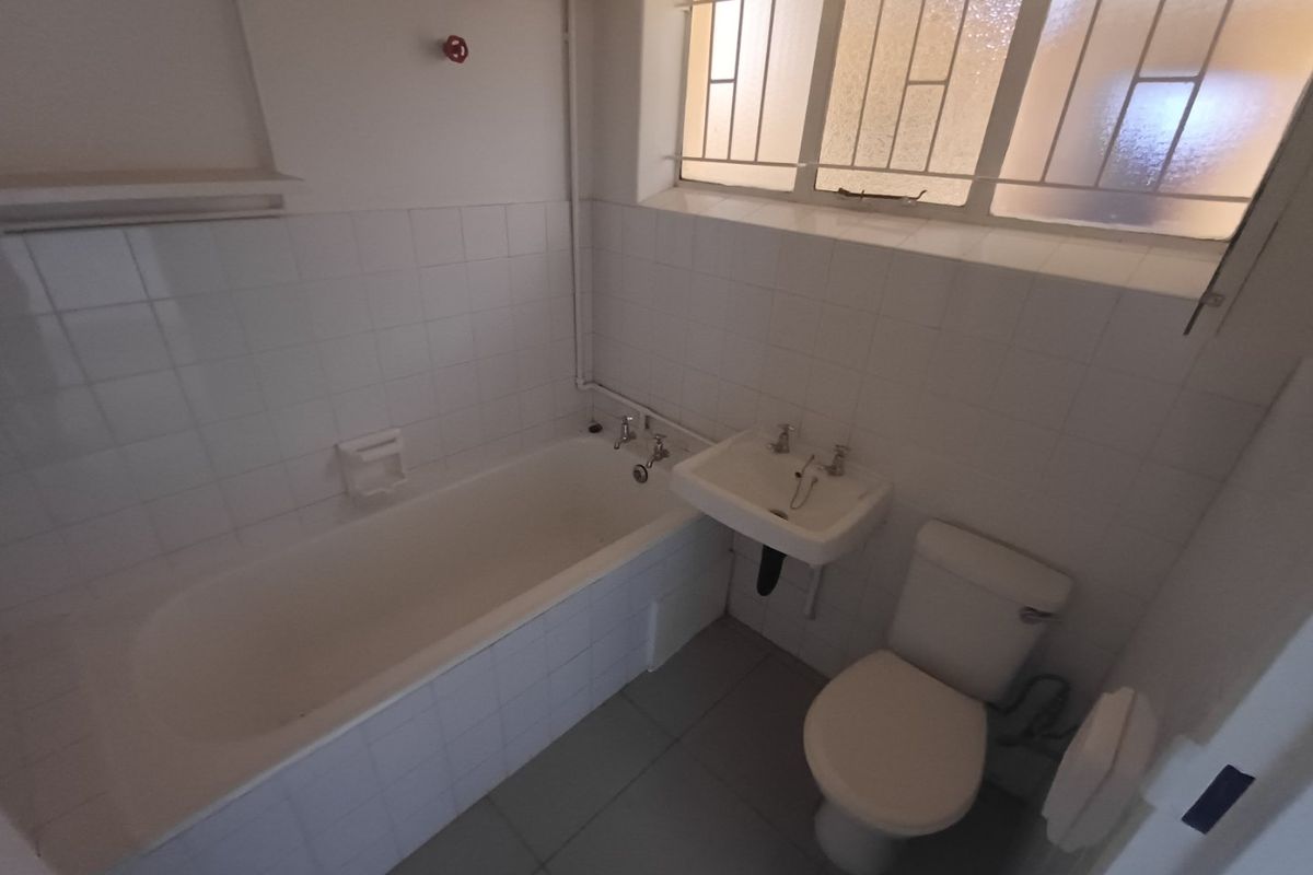 A functional bathroom featuring a standard bathtub, wall-mounted basin, and toilet, finished with white wall tiles and a window with security bars.