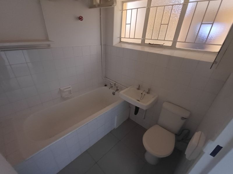 A functional bathroom featuring a standard bathtub, wall-mounted basin, and toilet, finished with white wall tiles and a window with security bars.
