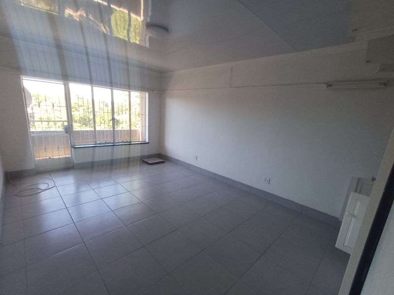 An empty, neutrally finished living area featuring large windows with security bars, tiled flooring, and a glossy ceiling finish.