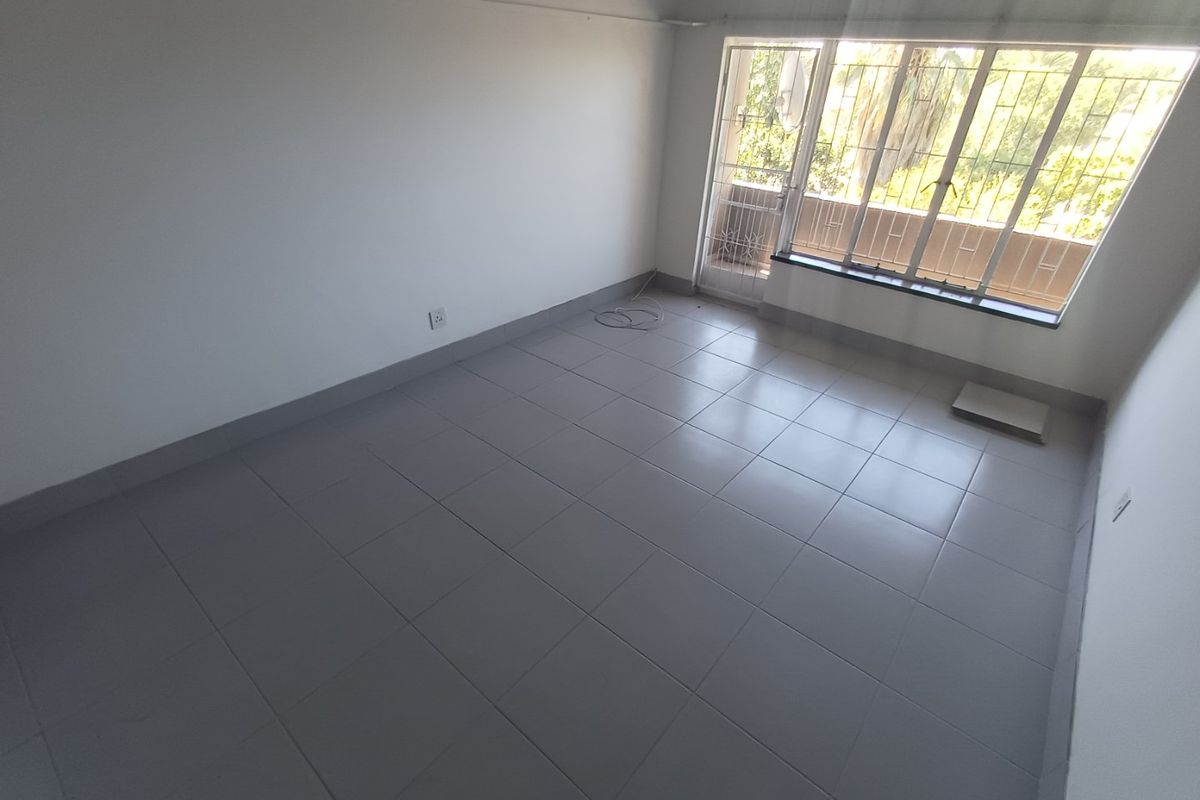 A bright, empty living room featuring neutral floor tiles, large windows with security bars, and access to an outdoor balcony area.