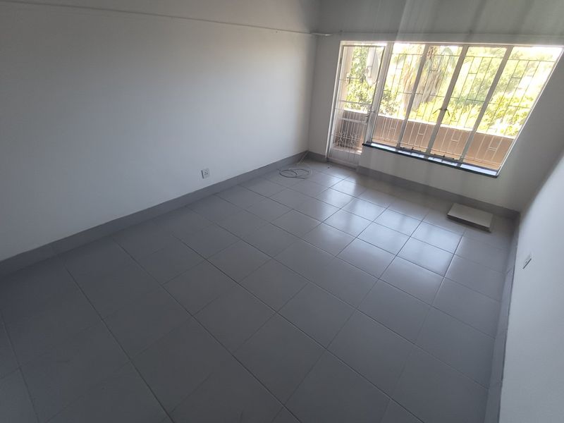 A bright, empty living room featuring neutral floor tiles, large windows with security bars, and access to an outdoor balcony area.