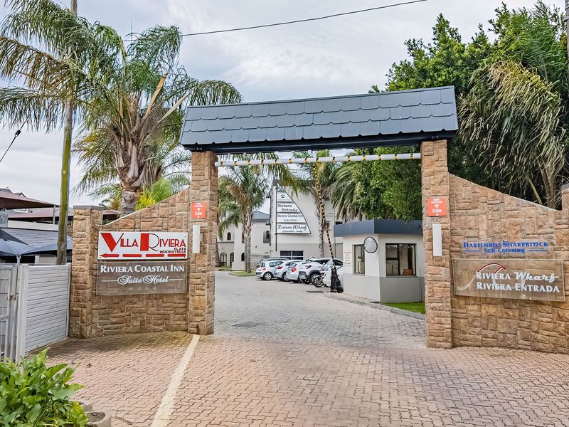 Main entrance gate to the Villa Riviera complex, featuring stone-clad pillars, a security boom gate, and a paved driveway leading into the property.