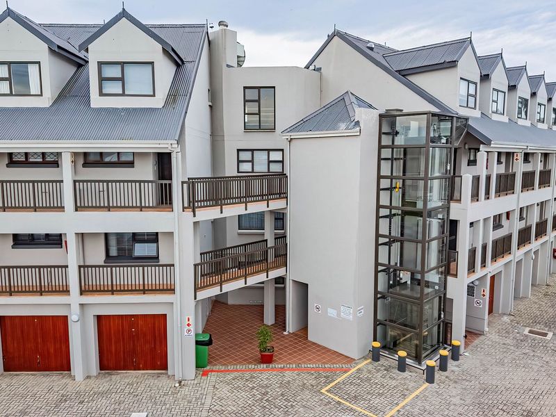 An elevated view of an apartment complex featuring a modern glass elevator, multiple balconies, and paved driveway access.