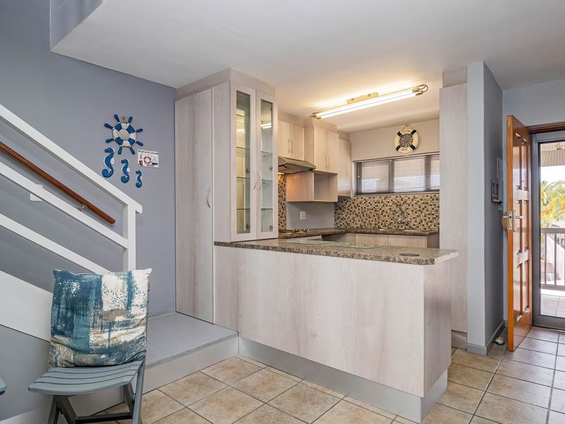 Compact kitchen featuring light wood-look cabinetry, granite countertops, and a mosaic tile backsplash, conveniently located adjacent to the staircase and exterior access door.