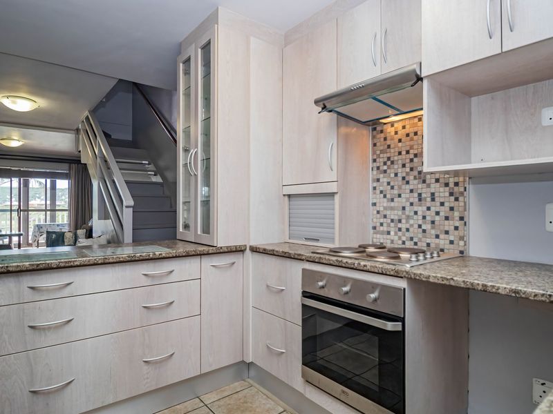 A functional kitchen featuring light wood-finish cabinetry, granite countertops, a mosaic tile backsplash, and integrated appliances, with an adjacent staircase leading to the living area.