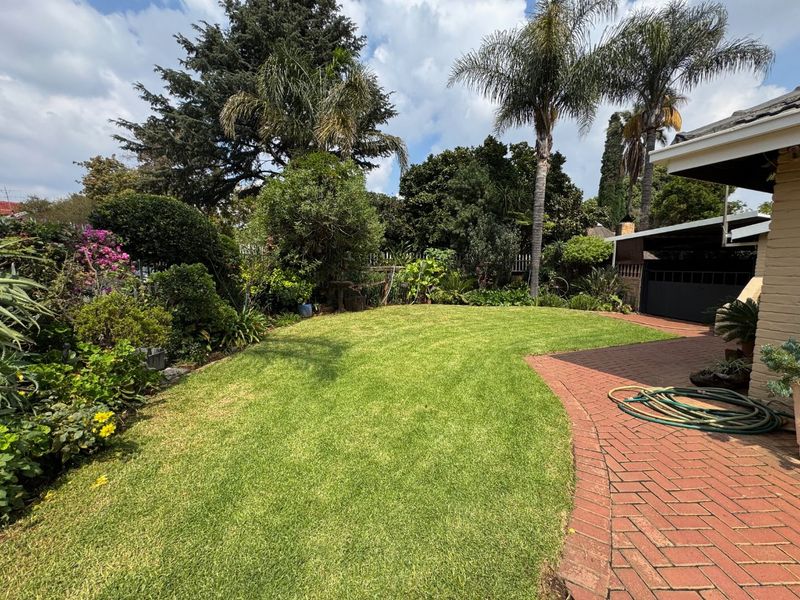 A well-maintained rear garden featuring a lush green lawn, mature trees, and brick-paved pathways leading to a covered patio area.